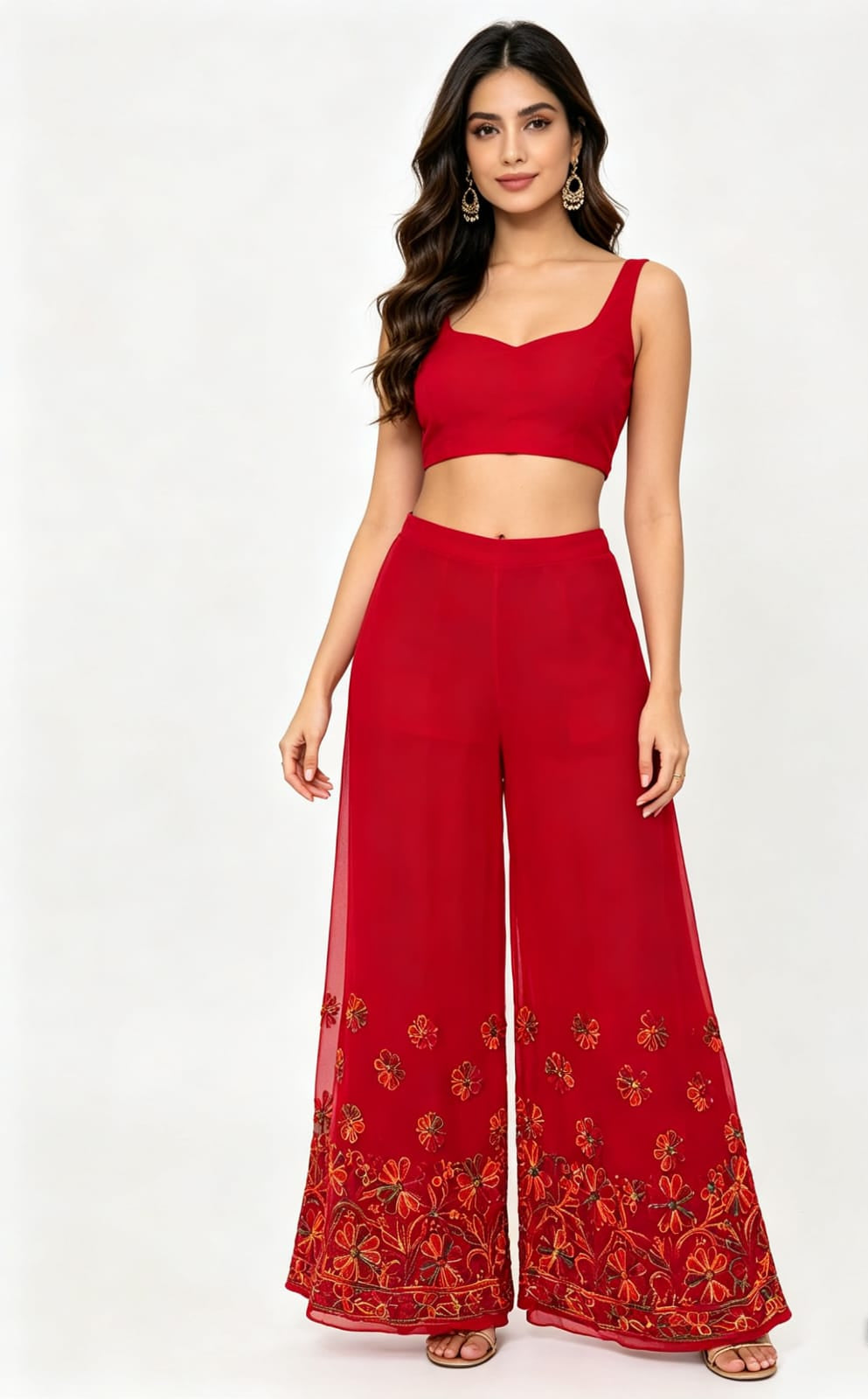 A close-up studio photograph focusing on the lower half of a woman wearing flowing, wide-leg red palazzo pants. The sheer fabric pants feature intricate, multi-colored floral and vine embroidery in shades of gold, orange, and green along the bottom hem and lower leg section. The upper part of her body and the top she is wearing are cropped out of the frame, making the embroidered palazzo pants the central focus. She is standing against a plain white background, and the lighting is even and bright.