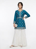 A model wearing a pair of pure white sharara pants featuring intricate Bakhiya embroidery and shimmering silver Kaamdani detailing, styled with a teal kurti against a plain white background.