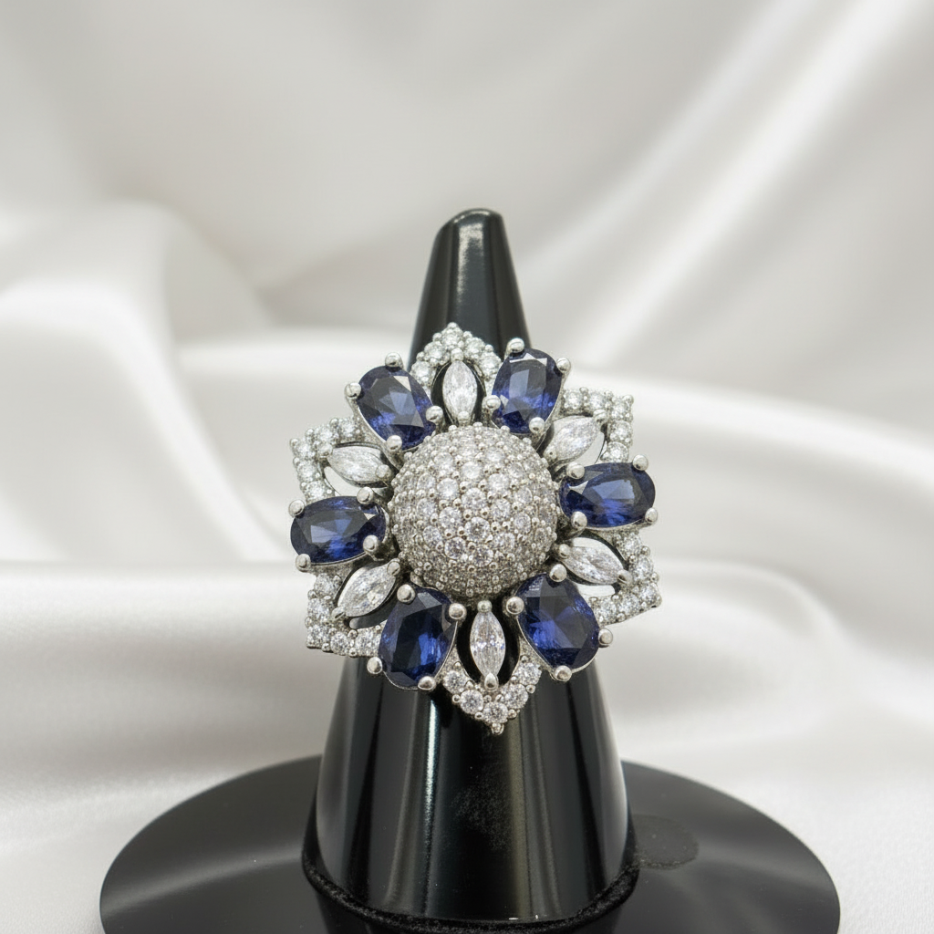 A floral-shaped silver ring brooch with deep blue oval-cut stones and clear pavé center and accents, displayed on a black cone stand against a gray gradient background. A white '2 cm' width measurement overlay is shown above the piece.