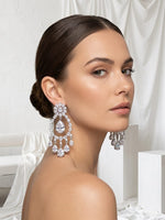 A woman with her hair in a low bun models a pair of large, sparkling silver-toned teardrop chandelier earrings with clear gemstones, set against a soft white draped fabric background.