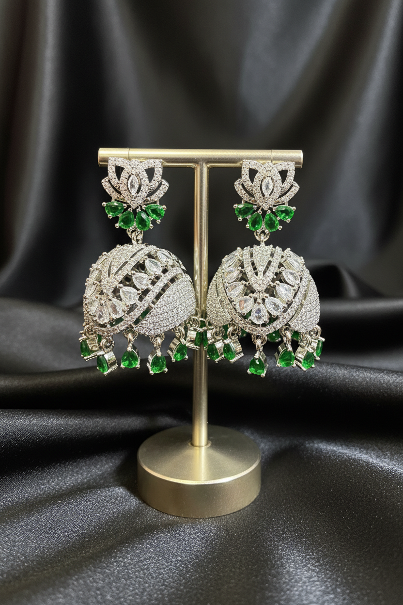 A pair of silver-toned Indian jhumka earrings with green crystal accents, displayed on a gold-colored stand against a dark textured background.