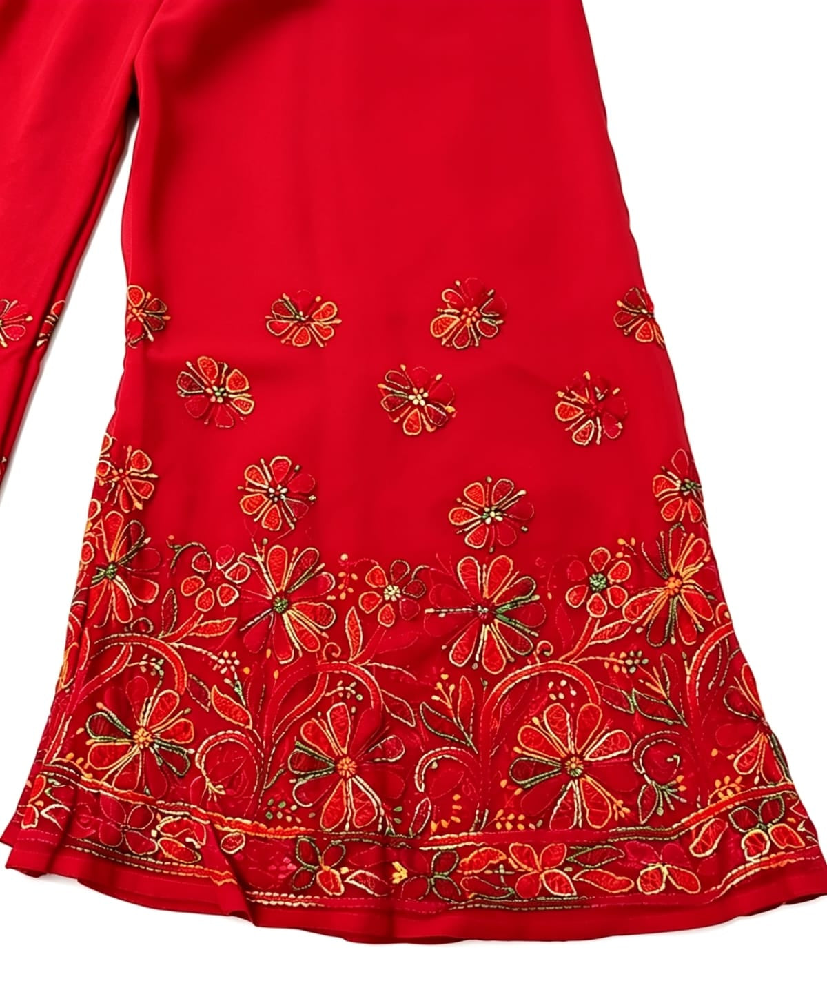 A close-up, flat-lay view of a red palazzo pant leg, featuring intricate, multi-color floral and vine embroidery at the hem and scattered motifs above. Against a plain white background.