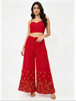 A close-up studio photograph focusing on the lower half of a woman wearing flowing, wide-leg red palazzo pants. The sheer fabric pants feature intricate, multi-colored floral and vine embroidery in shades of gold, orange, and green along the bottom hem and lower leg section. The upper part of her body and the top she is wearing are cropped out of the frame, making the embroidered palazzo pants the central focus. She is standing against a plain white background, and the lighting is even and bright.