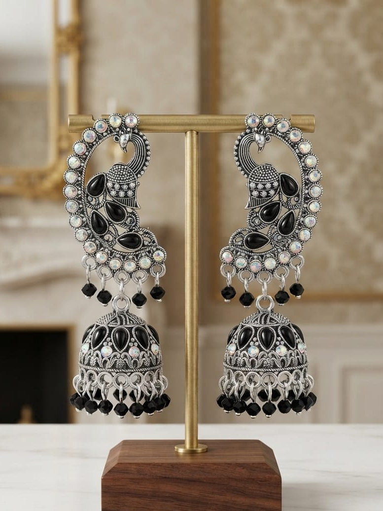 A pair of ornate silver-toned Indian jhumka earrings with black stones and white pearl-like accents, displayed on a gold stand with a wooden base on a marble surface.