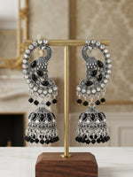 A pair of ornate silver-toned Indian jhumka earrings with black stones and white pearl-like accents, displayed on a gold stand with a wooden base on a marble surface.