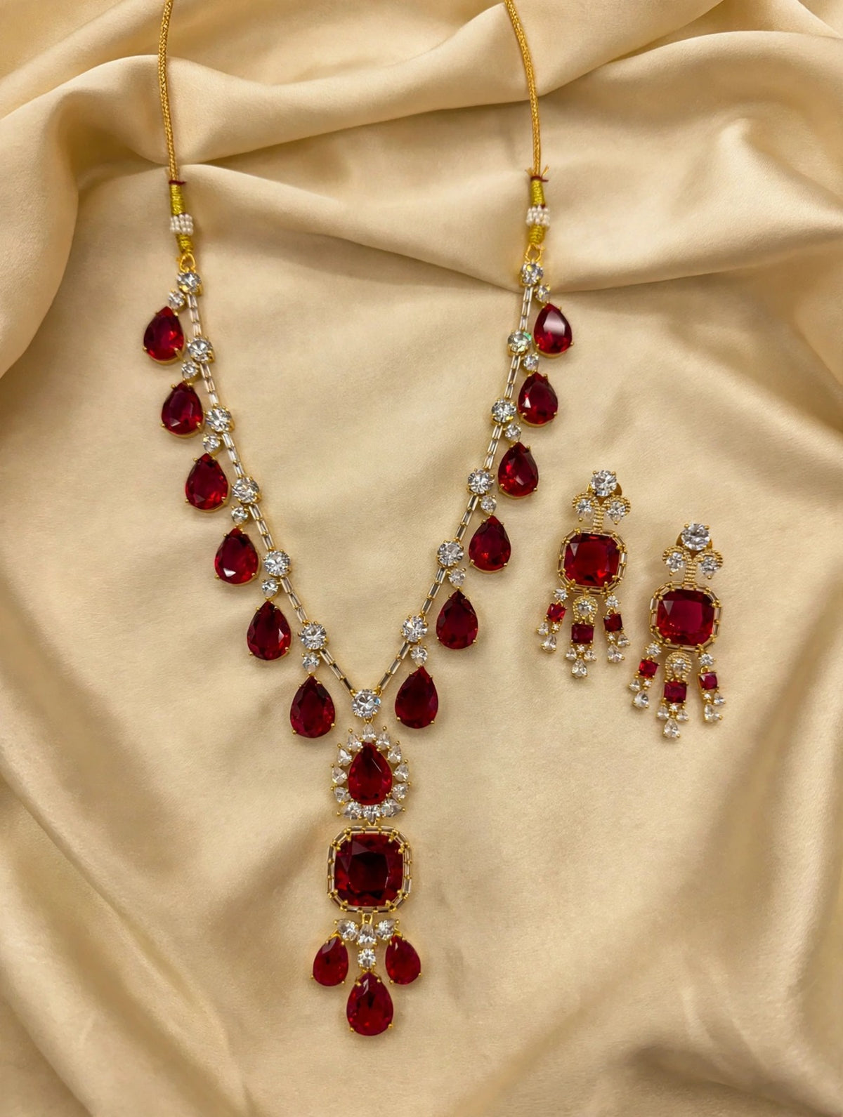 Ruby red pear & cushion-cut gemstone necklace with matching statement earrings, gold-tone finish with sparkling clear crystal accents — South Asian bridal jewelry laid on cream silk fabric.