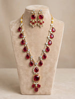 Ruby red pear & cushion-cut gemstone necklace with matching statement earrings, gold-tone finish with sparkling clear crystal accents — South Asian bridal jewelry displayed on beige velvet bust.