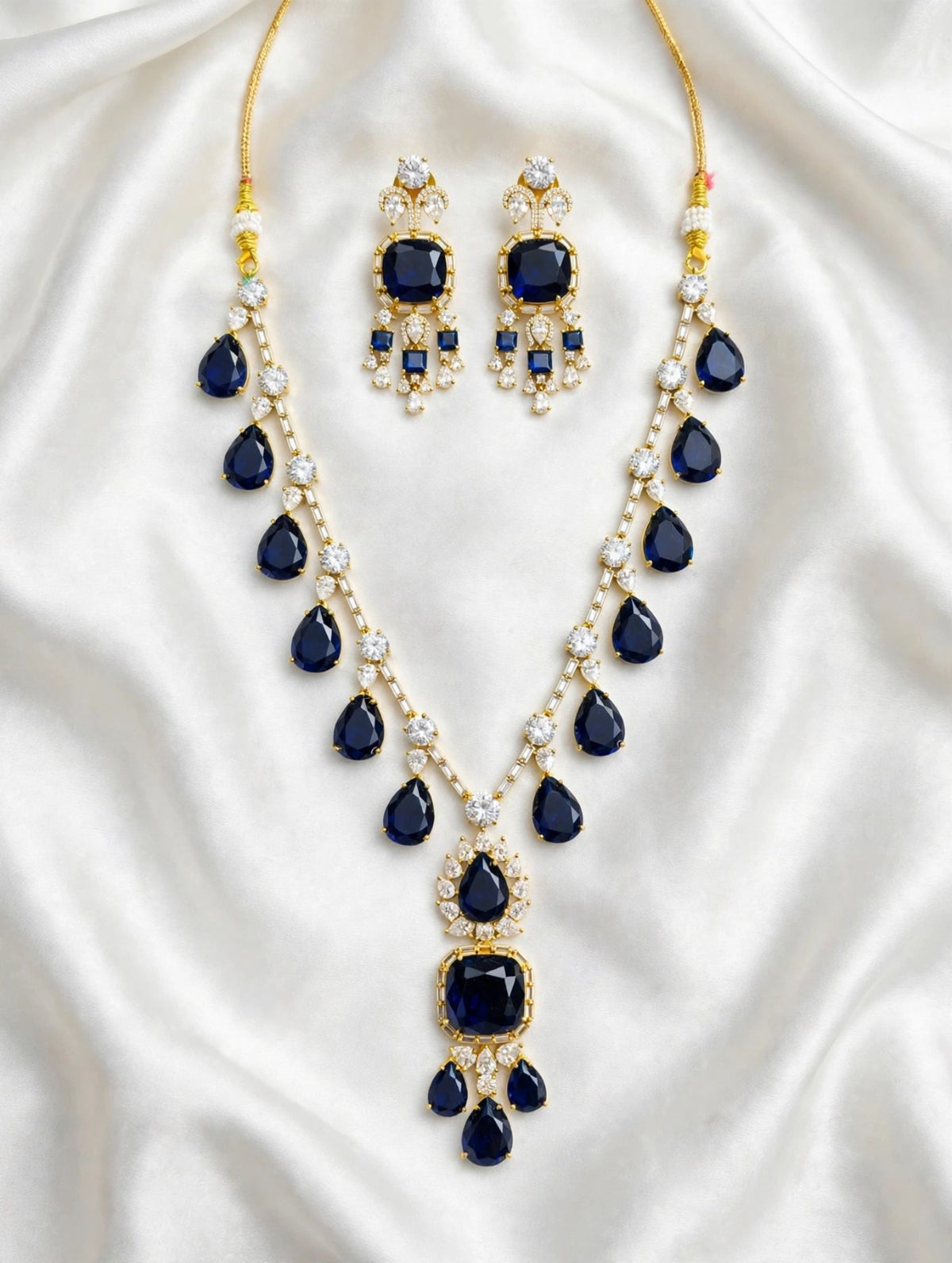 Sapphire blue pear & cushion-cut gemstone necklace with matching statement earrings, gold-tone finish with sparkling clear crystal accents — South Asian bridal & formal jewelry set on white silk background.