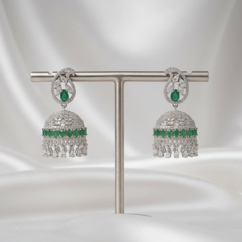 A pair of silver-toned jhumka earrings with green stone accents, displayed on a silver T-stand against a dark gray background.