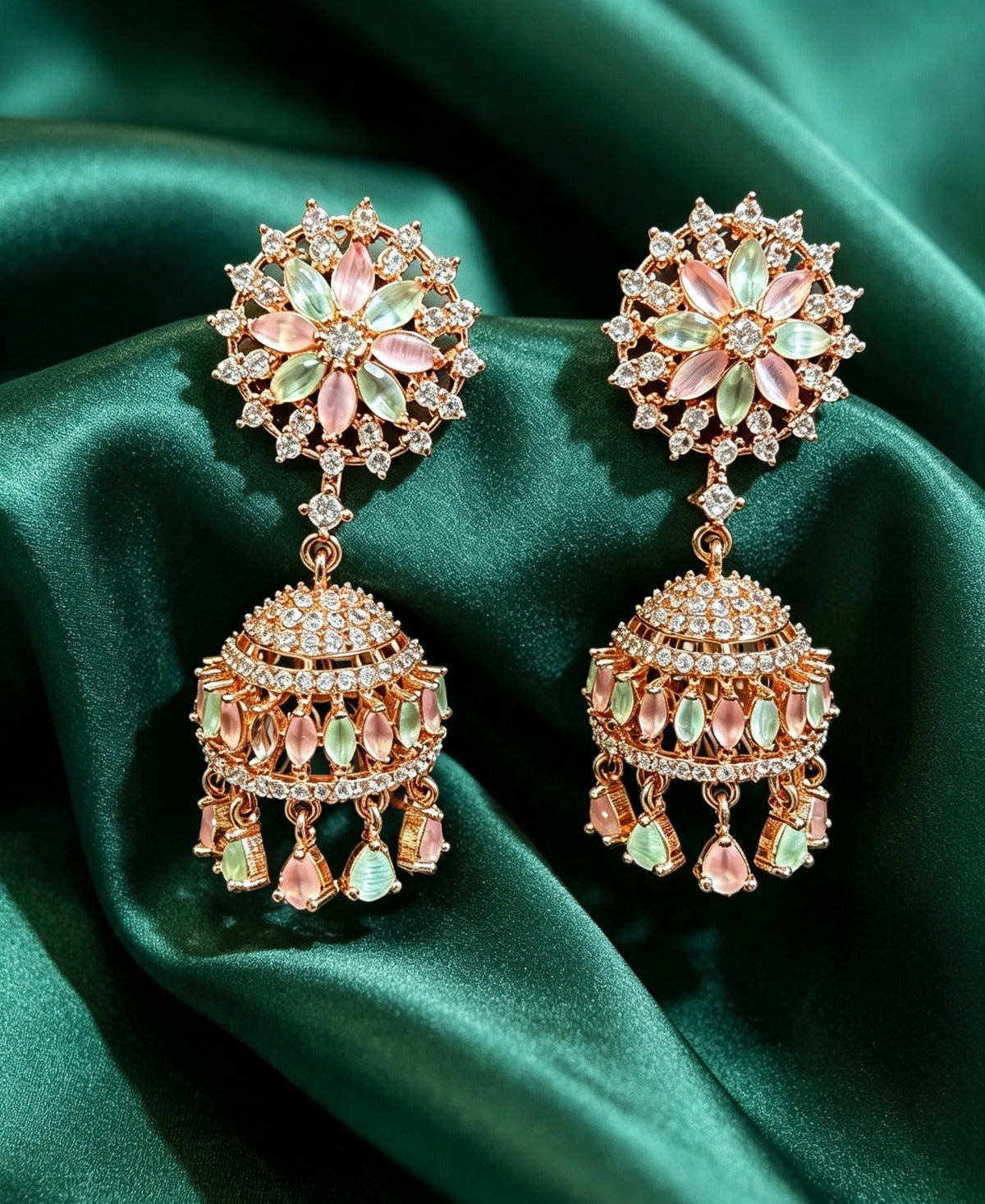 Luxurious rose gold-plated floral CZ jhumka earrings with pastel pink and mint green stones displayed on rich emerald green silk fabric.