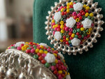 A close-up of a decorative brooch featuring a circular silver-tone beaded border and a center filled with multicolored seed beads, white rose-shaped beads, and small white flower charms, displayed on a dark green velvet surface.