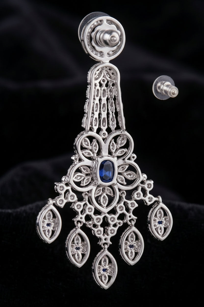 A close-up view of the back of an elegant chandelier earring featuring a central blue sapphire stone, intricate filigree metalwork, and sparkling diamond accents, shown with a clear rubber earring back.