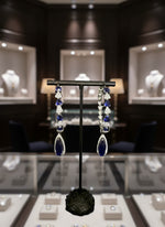 A pair of silver finish American diamond Jhumka bali earrings with green and clear stones on a stand.