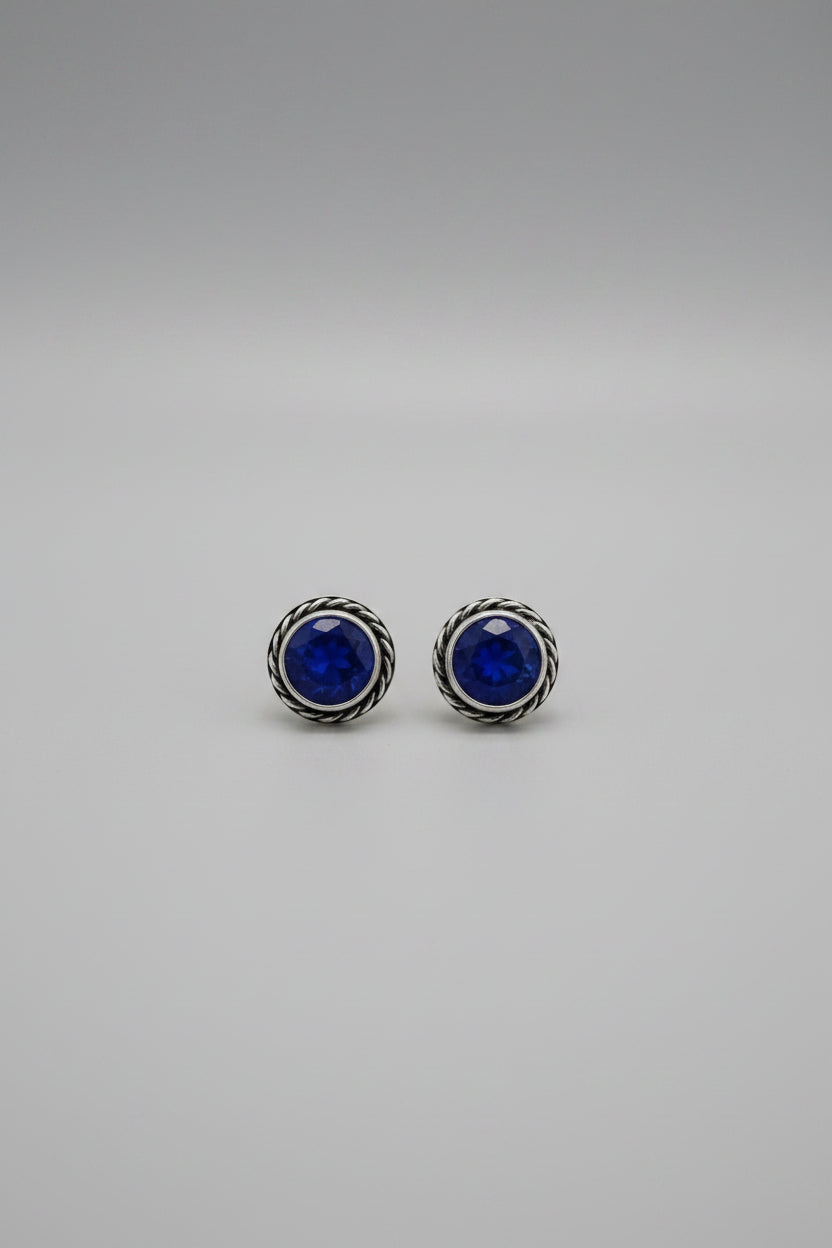 A pair of round stud earrings with deep blue gemstones and silver-toned twisted bezel settings, resting on a light wood tabletop.
