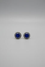 A pair of round stud earrings with deep blue gemstones and silver-toned twisted bezel settings, resting on a light wood tabletop.