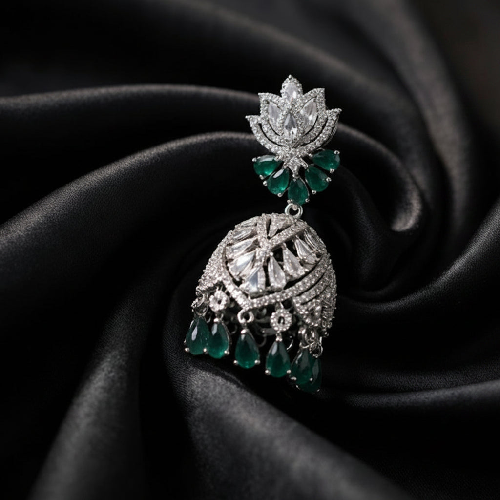 A silver-toned traditional Indian jhumka earring with green emerald-cut gemstone accents, displayed on a dark black satin fabric background.