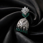 A silver-toned traditional Indian jhumka earring with green emerald-cut gemstone accents, displayed on a dark black satin fabric background.