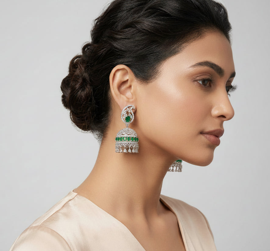 A woman in profile wearing a pair of silver-toned jhumka earrings with green stone accents and a delicate silver chain necklace, set against a clean white background.