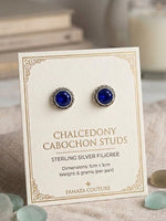 A pair of cabochon stud earrings with deep blue stones and silver filigree settings, mounted on a cream display card. The card is labeled 'CHALCEDONY CABOCHON STUDS' and shows the brand 'FAMAZA COUTURE'.