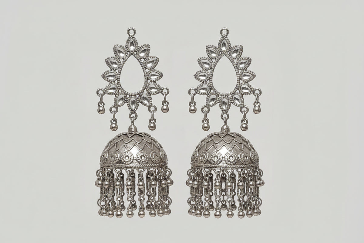 A close-up of a model's ear wearing a large, ornate silver-toned jhumka earring with intricate filigree detailing and dangling bell-shaped charms.