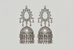 A close-up of a model's ear wearing a large, ornate silver-toned jhumka earring with intricate filigree detailing and dangling bell-shaped charms.