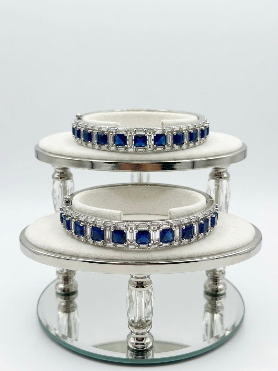 Pair of silver-plated hinged bangles featuring deep sapphire blue square stones and clear baguette-cut American Diamonds on a luxury display stand.