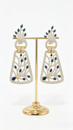 Pair of silver-toned dangle earrings with royal blue stone embellishments and American Diamond lattice detailing on a posh display stand.