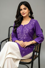 Seated view of a woman wearing a royal purple cotton short kurti featuring intricate white floral embroidery on the yoke and front, a keyhole neckline, and a square hem line, paired with white trousers.