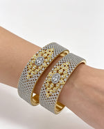 A model wearing a pair of luxury wide-band dual-tone bangles, featuring a full pavé American Diamond surface with an intricate gold floral centerpiece.