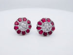 A close-up product photo of a pair of silver floral cluster earrings featuring vibrant ruby-red pear-shaped stone petals surrounding a center cluster of sparkling clear marquise and round stones, resting side-by-side on a light marble surface.
