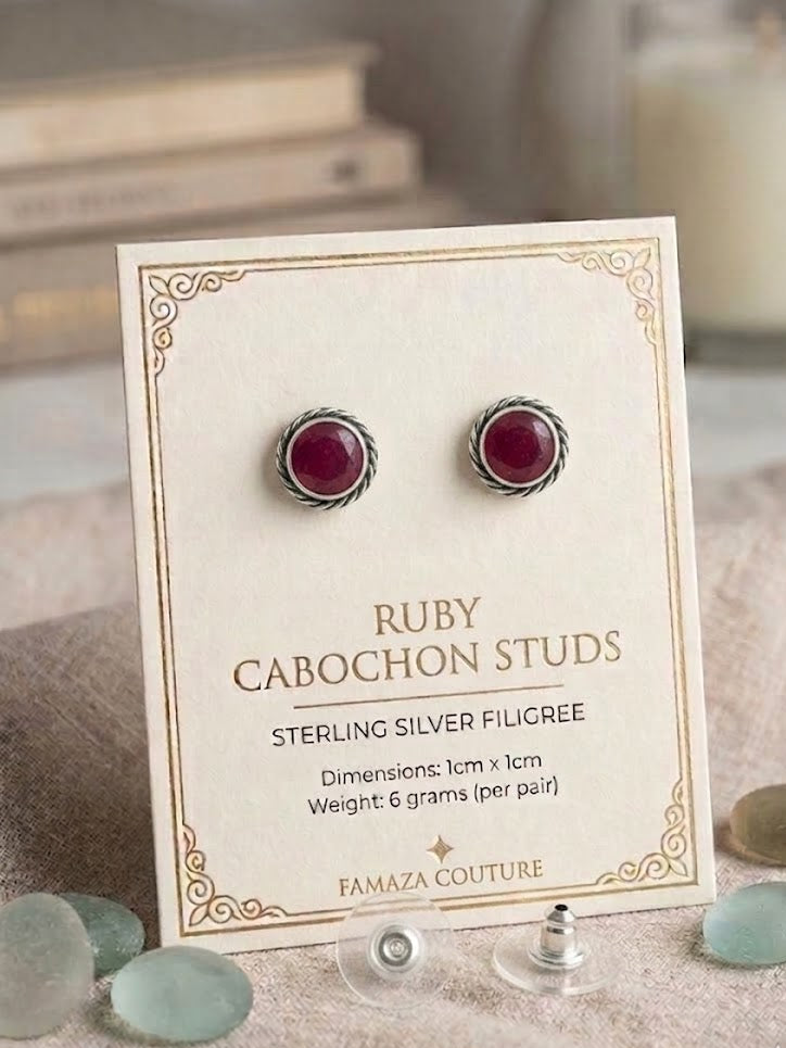 Ruby cabochon stud earrings in sterling silver on a cream display card, with the text 'RUBY CABOCHON STUDS' and 'FAMAZA COUTURE' visible. The card is styled on a fabric surface with sea glass and a blurred candle in the background.