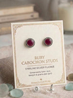 Ruby cabochon stud earrings in sterling silver on a cream display card, with the text 'RUBY CABOCHON STUDS' and 'FAMAZA COUTURE' visible. The card is styled on a fabric surface with sea glass and a blurred candle in the background.