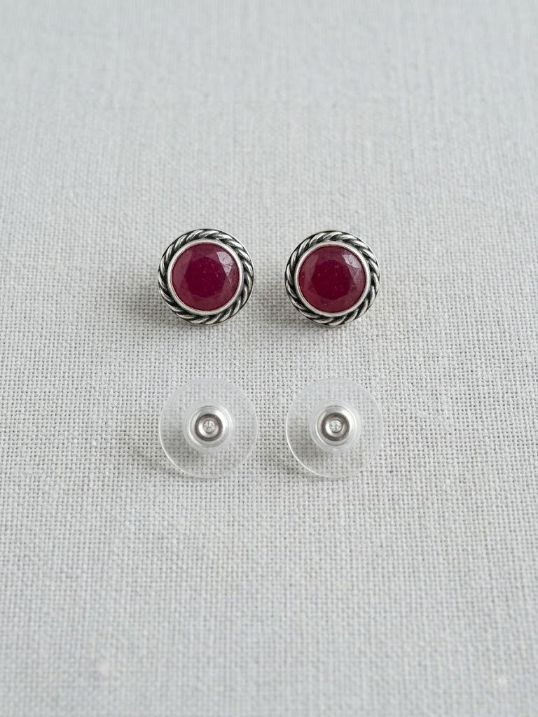 A pair of silver-toned round stud earrings with deep red gemstone centers, shown above their clear plastic backing clips on a light gray textured fabric background.
