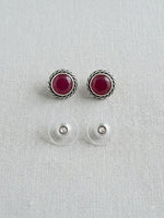 A pair of silver-toned round stud earrings with deep red gemstone centers, shown above their clear plastic backing clips on a light gray textured fabric background.