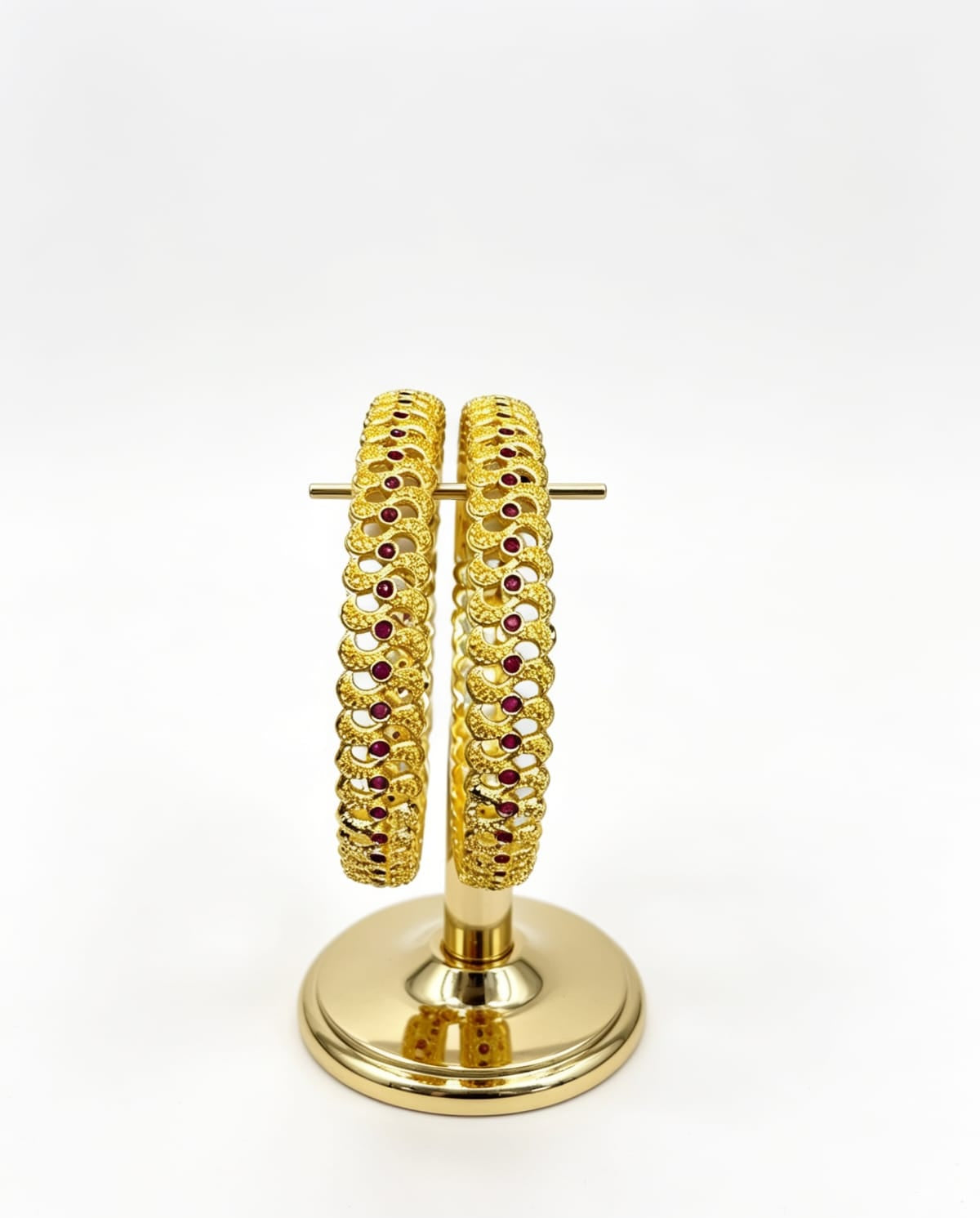A pair of gold-plated bangles with an intricate scalloped texture and small ruby red stones set on a gold display stand.