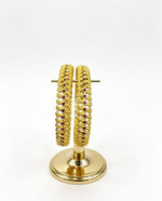 A pair of gold-plated bangles with an intricate scalloped texture and small ruby red stones set on a gold display stand.