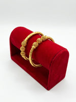A pair of micro gold-plated copper bangles featuring three intricate hollow filigree orbs and a textured band, displayed on a posh red velvet jewelry stand against a pure white background.