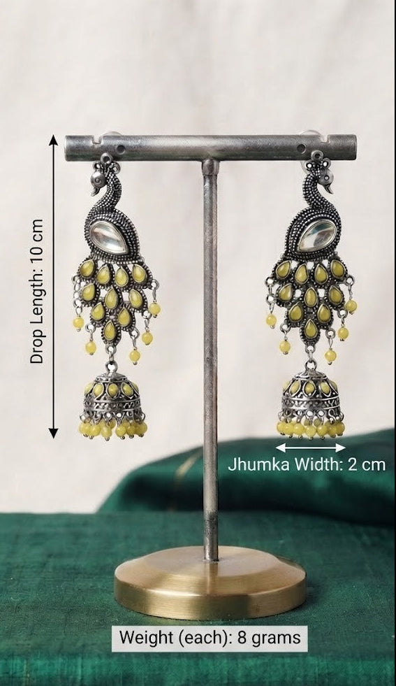 Silver-toned peacock jhumka earrings with yellow bead drops, displayed on a metal stand with measurement labels indicating drop length 10 cm, jhumka width 2 cm, and weight 8 grams each.