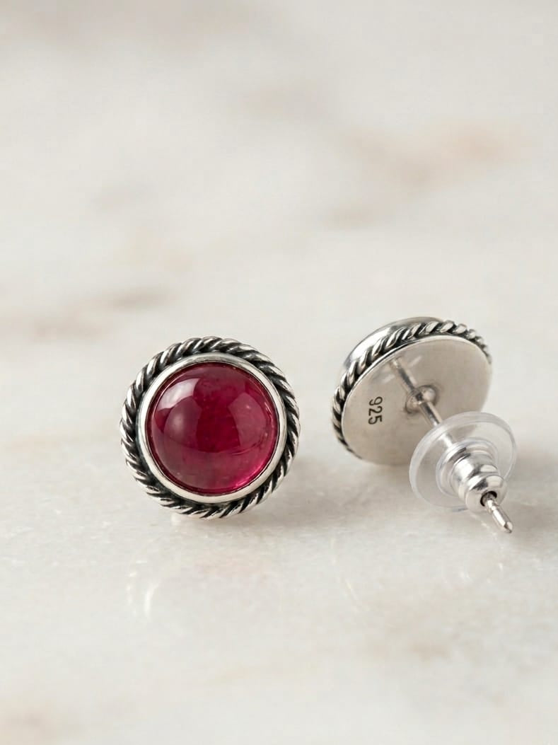 A pair of sterling silver stud earrings with round deep red gemstone centers and a twisted rope border, displayed on a light marble surface. One earring is shown from the front, and the other is angled to show the back with a clear protective cap and a '925' silver stamp.
