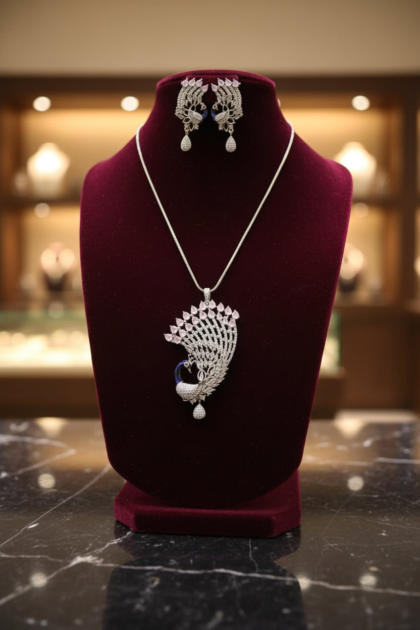 Silver plated a set of peacock-shaped pendant in silver chain with peacock-shaped earrings. The pendant is detailed with blue enamel and has sparkling crystal pink and white AD stones embellishments. Party wear jewellery set. Jewellery set is displayed on a black velvet jewellery dummy.