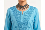 A close-up view of a person wearing a light blue short kurti featuring intricate tonal floral embroidery around the split round neckline, styled with an ornate silver-toned choker necklace.