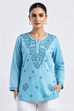 A front-facing portrait of a model wearing a sky blue short kurti with detailed tonal embroidery along the split neckline and scattered floral motifs across the front, paired with white trousers.