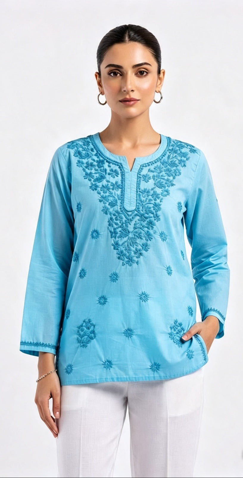 A model wearing a light sky blue short kurti with intricate tonal embroidery around the split neckline and scattered floral motifs across the front, paired with white trousers.