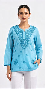 A model wearing a light sky blue short kurti with intricate tonal embroidery around the split neckline and scattered floral motifs across the front, paired with white trousers.
