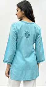 A model shown from the back, wearing a sky blue cotton short kurti with matching embroidery around the neckline and a floral motif on the upper back, against a white studio background.