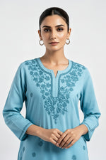 A model with arms crossed wearing a light blue short kurti featuring intricate tonal embroidery along the split neckline and sleeves.