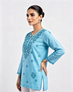 A side profile view of a model wearing a light blue short kurti featuring tonal floral embroidery, side slits, and embroidered sleeve cuffs, paired with white trousers.