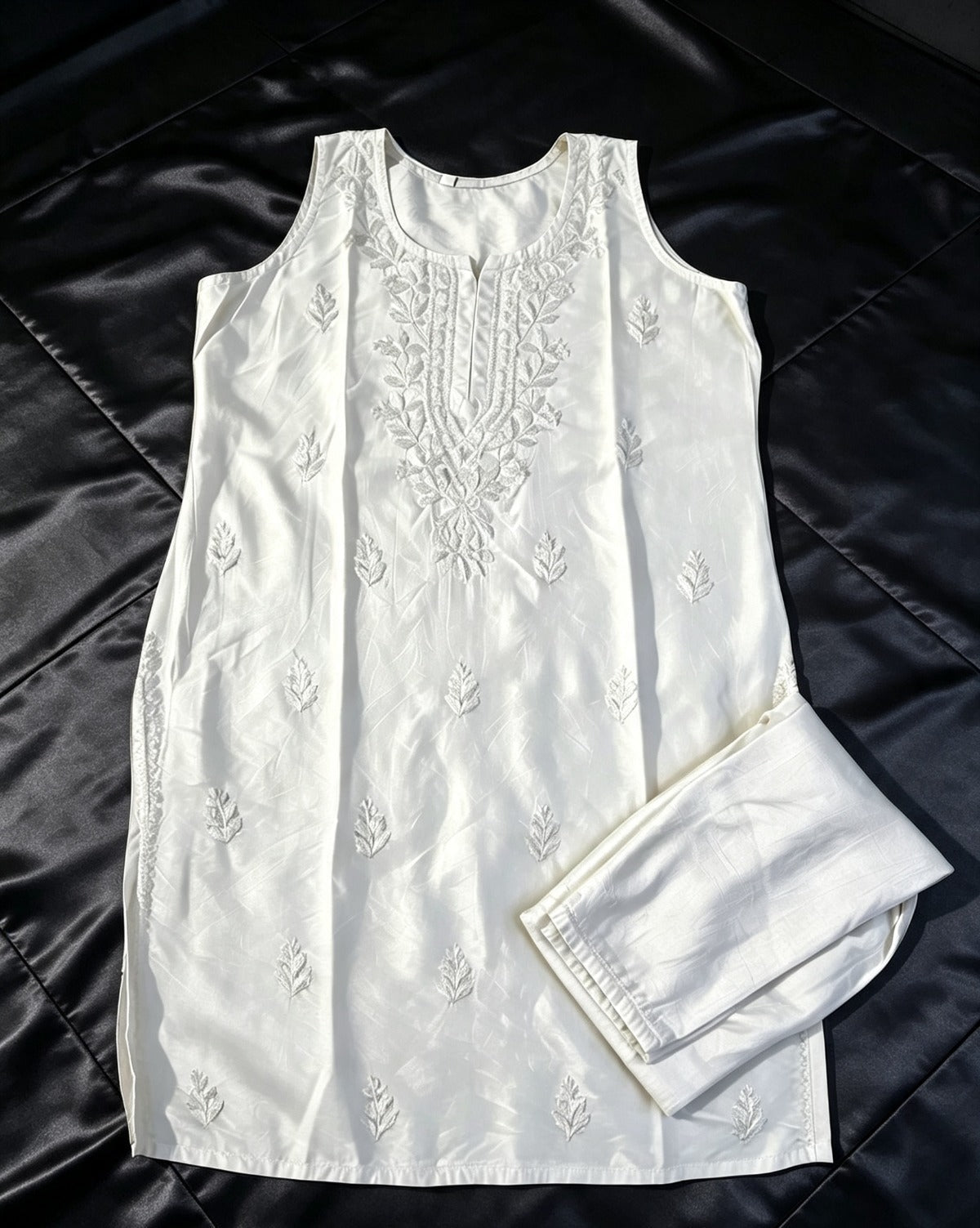 Flat lay view of the white handcrafted sleeveless Chanderi silk Chikankari tunic and matching folded trousers arranged on a black satin surface.