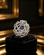 Exquisite Silver & Blue statement ring featuring a square-cut blue centre stone, intricate layered geometric diamond halo, and polished silver finish — elegantly displayed on a premium black velvet jewellery stand with golden lighting.
