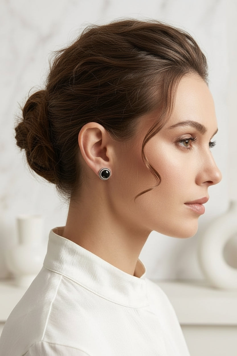 A woman in profile wearing a small round stud earring with a black center and a silver outer ring, against a soft light gray marble background.